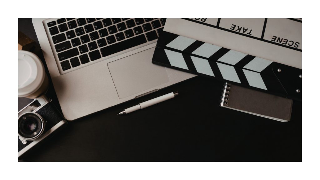 un ordinateur ouvert vu sur le clavier, un appareil photo, un clap de cinema, un ca;hier fermé et un casque. 